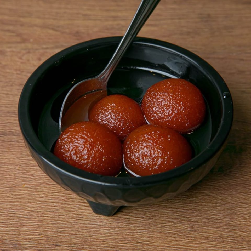 Four glistening gulab jamuns in a black bowl with a spoon, on a wooden table.