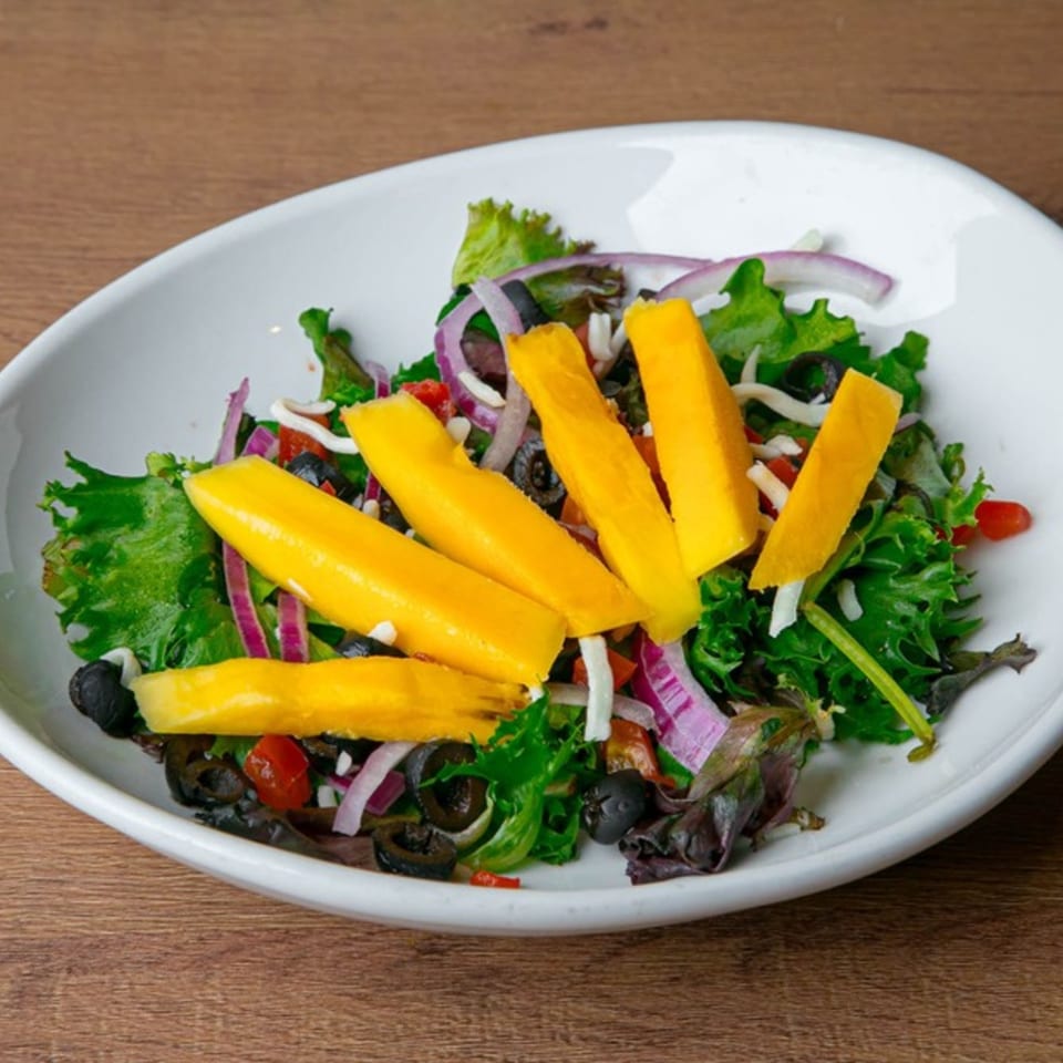 Fresh green salad with mango slices, red onion, black olives, and cherry tomatoes in a white bowl.