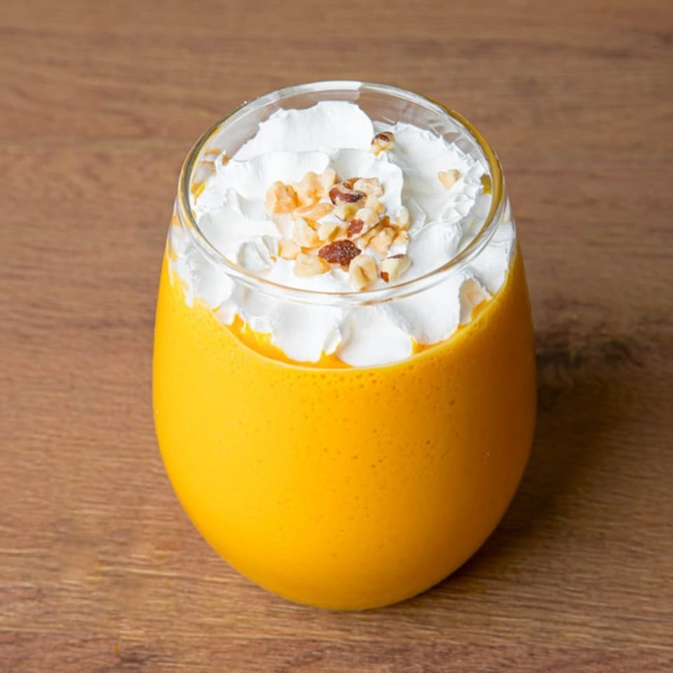 A glass of bright orange mango lassi topped with white whipped cream and chopped nuts.