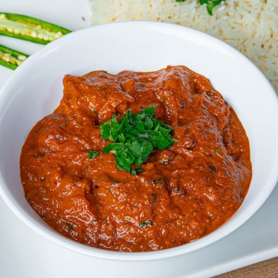 A white bowl filled with vibrant red Indian curry, garnished with fresh green herbs, alongside rice and chilies.